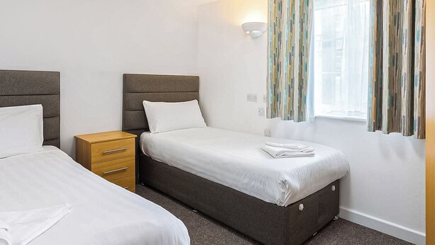 A hotel room with two beds, a wooden nightstand, a wall-mounted light, and a window with curtains. - 5G Larkstone - UKC9000 (Illfracombe)