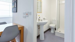 Ein Zimmer mit Schreibtisch, Stuhl, weißen Wänden, Waschbecken, Toilette und Dusche.
