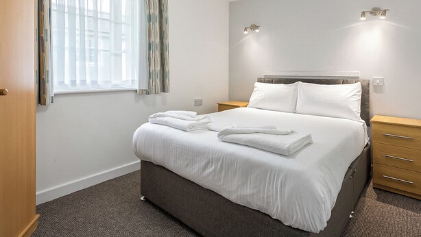 A hotel room with a bed, a nightstand, a wooden dresser, a window with curtains, and wall-mounted lighting.