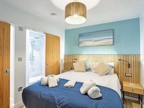 A bedroom with a bed, wooden headboard, a painting, a nightstand, and a bathroom with a shower. - 178 Residences - UKC8998 (Illfracombe)
