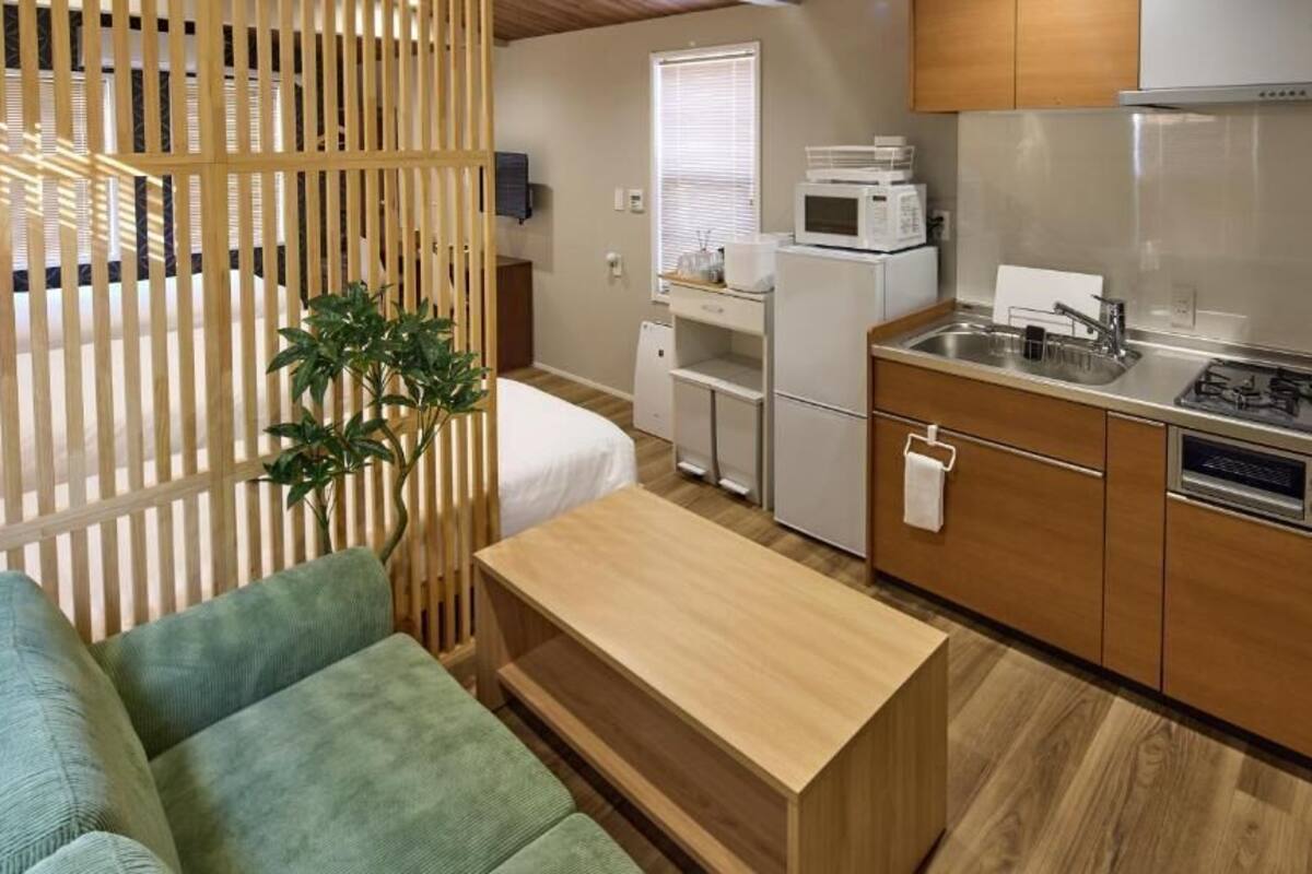A compact kitchenette with a microwave, stove, and sink. There is a small refrigerator and a wooden dining table. A green couch is visible in the foreground.