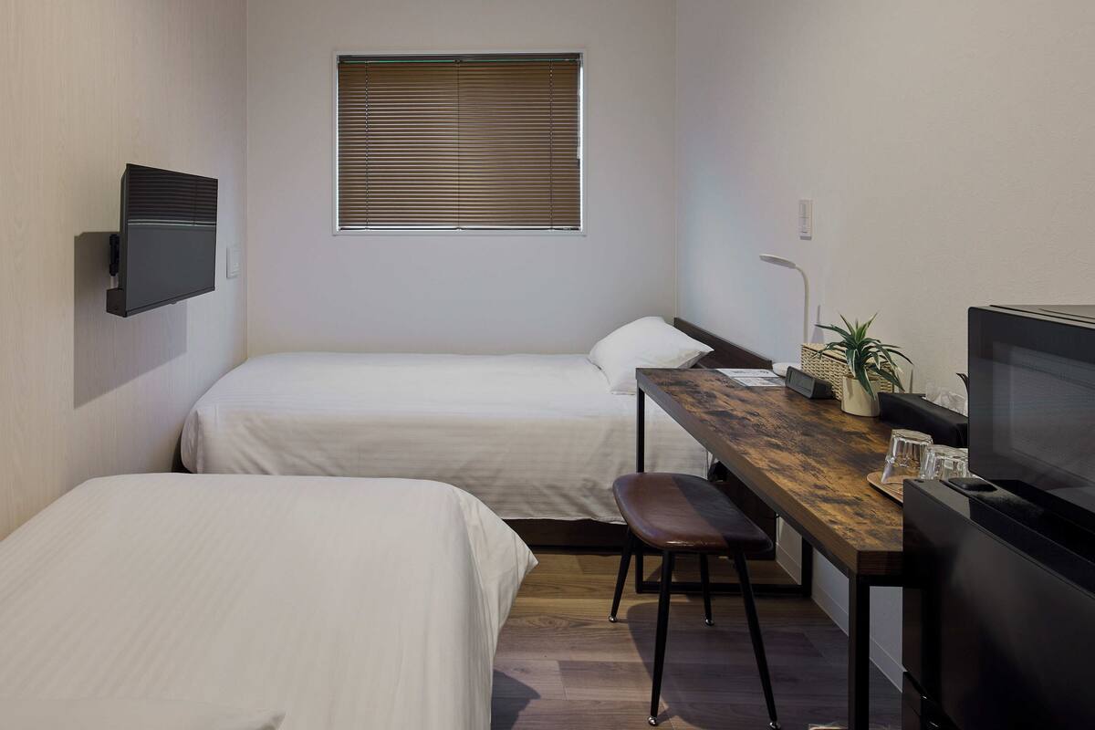 A small, modern hotel room with two single beds, a desk with a computer, a TV, and a window with blinds.