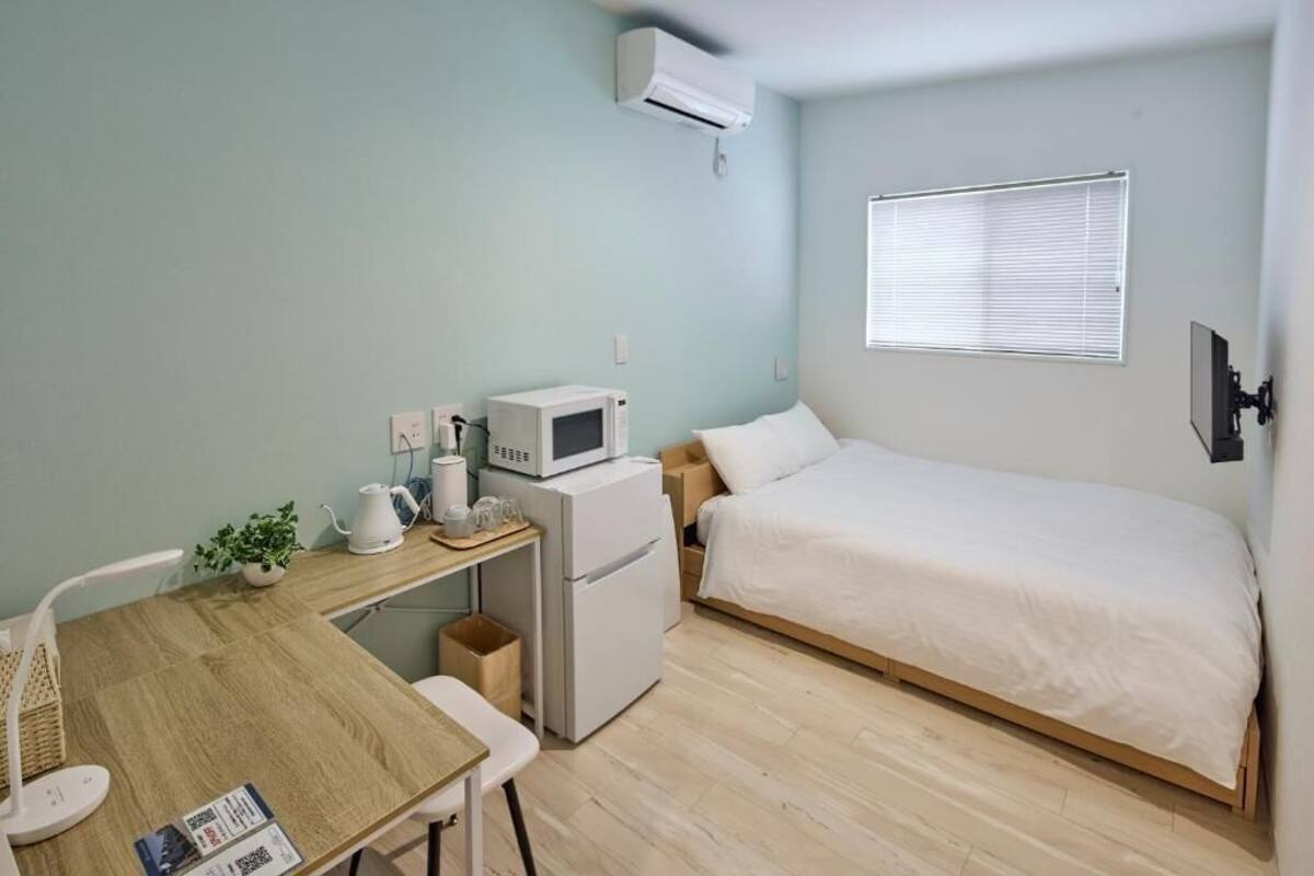 A compact bedroom with a bed, a small desk with a microwave, a refrigerator, a window with blinds, and a wall-mounted TV.