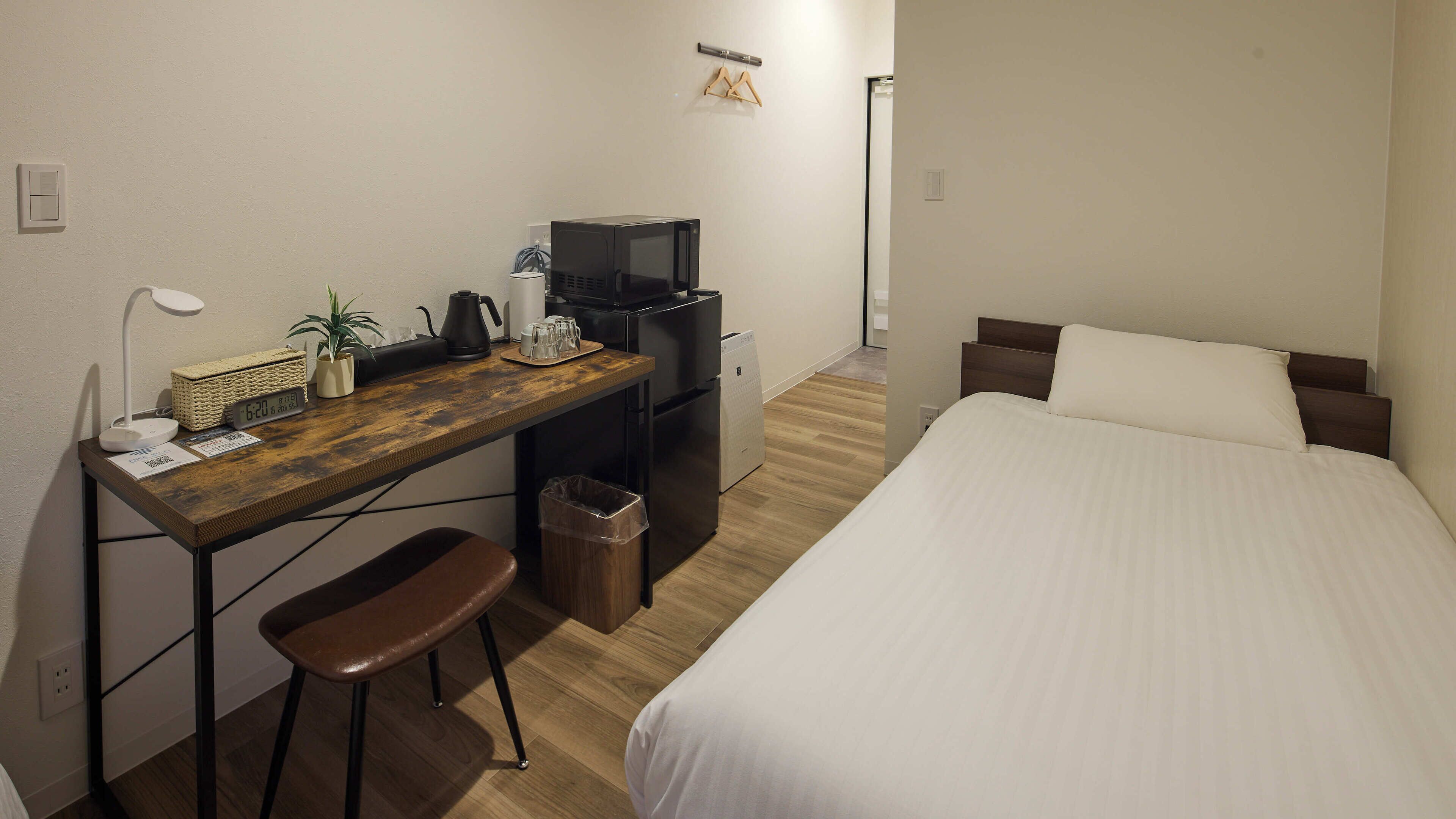 Ein kleines Hotelzimmer mit einem Holztisch, einem kleinen Hocker, einem Bett mit weißer Bettwäsche, einem schwarzen Kühlschrank und einem an der Wand montierten Fernseher.