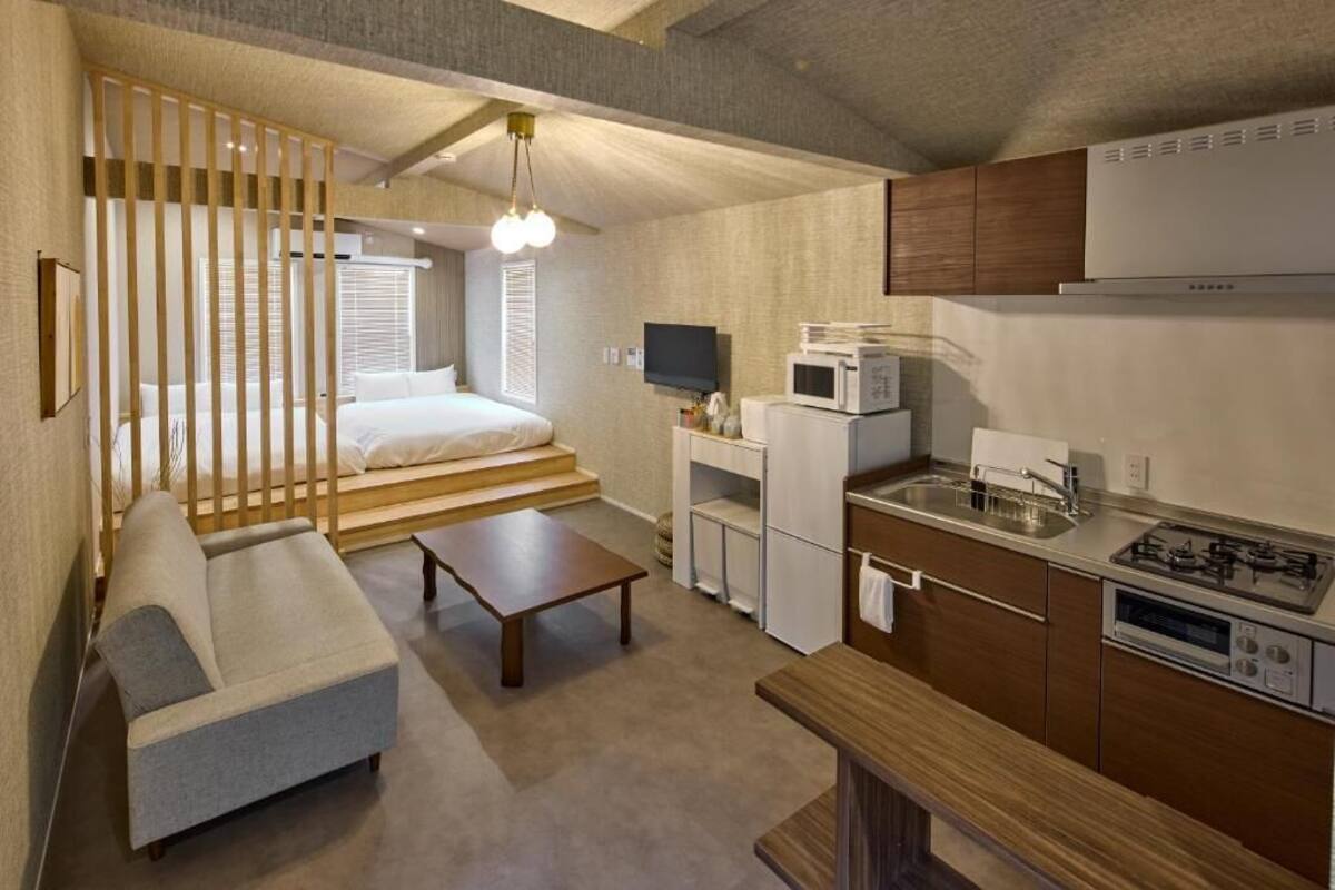 A compact, modern room with a sofa, a small table, a kitchenette with a stove and microwave, and a bed with a view of the outdoors.