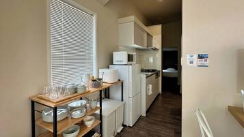 A compact kitchen with white appliances, a wooden shelf with dishes, and a window with blinds.