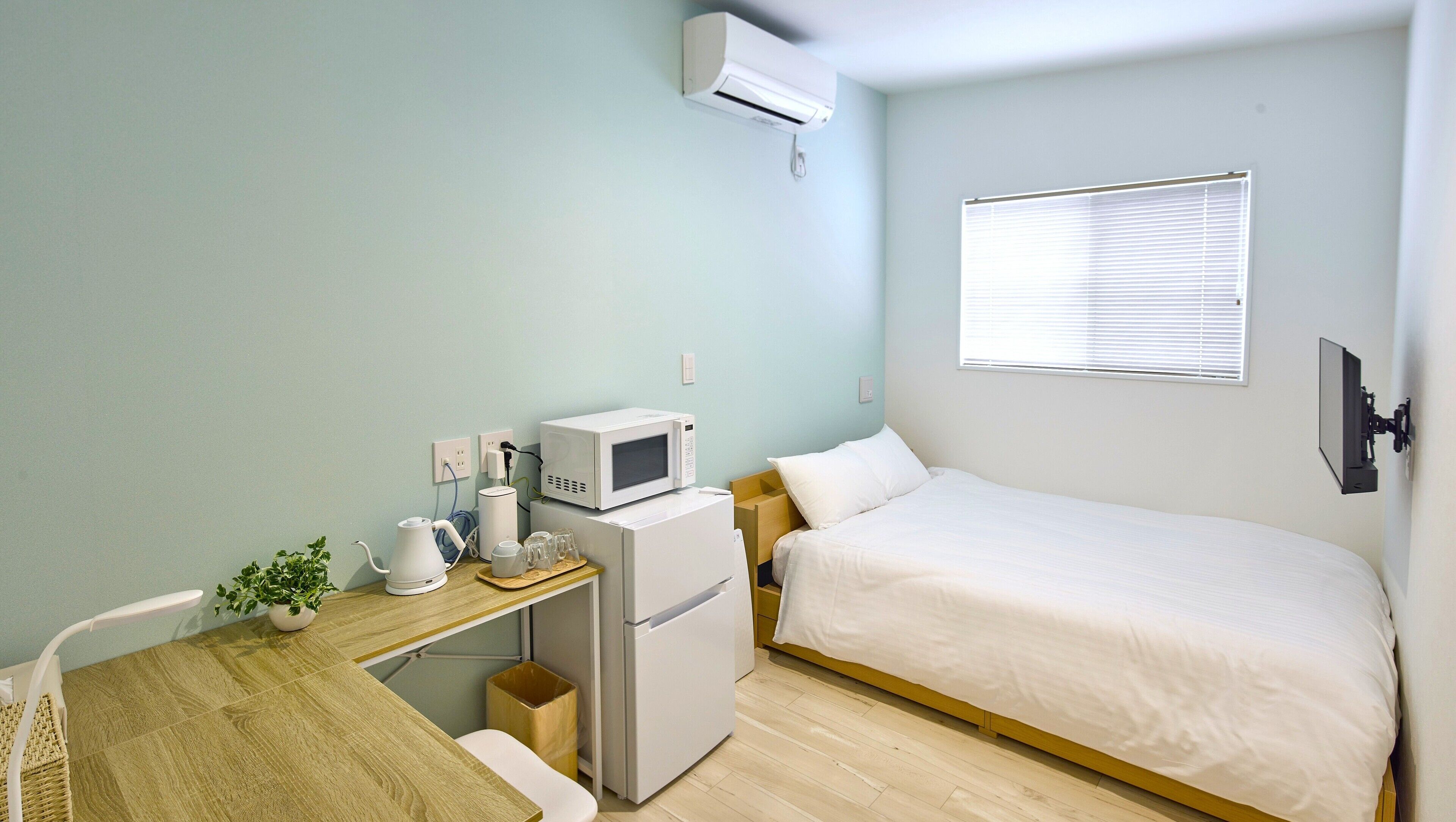 A small, clean room with a bed, a desk with a microwave, a refrigerator, and a window with blinds.