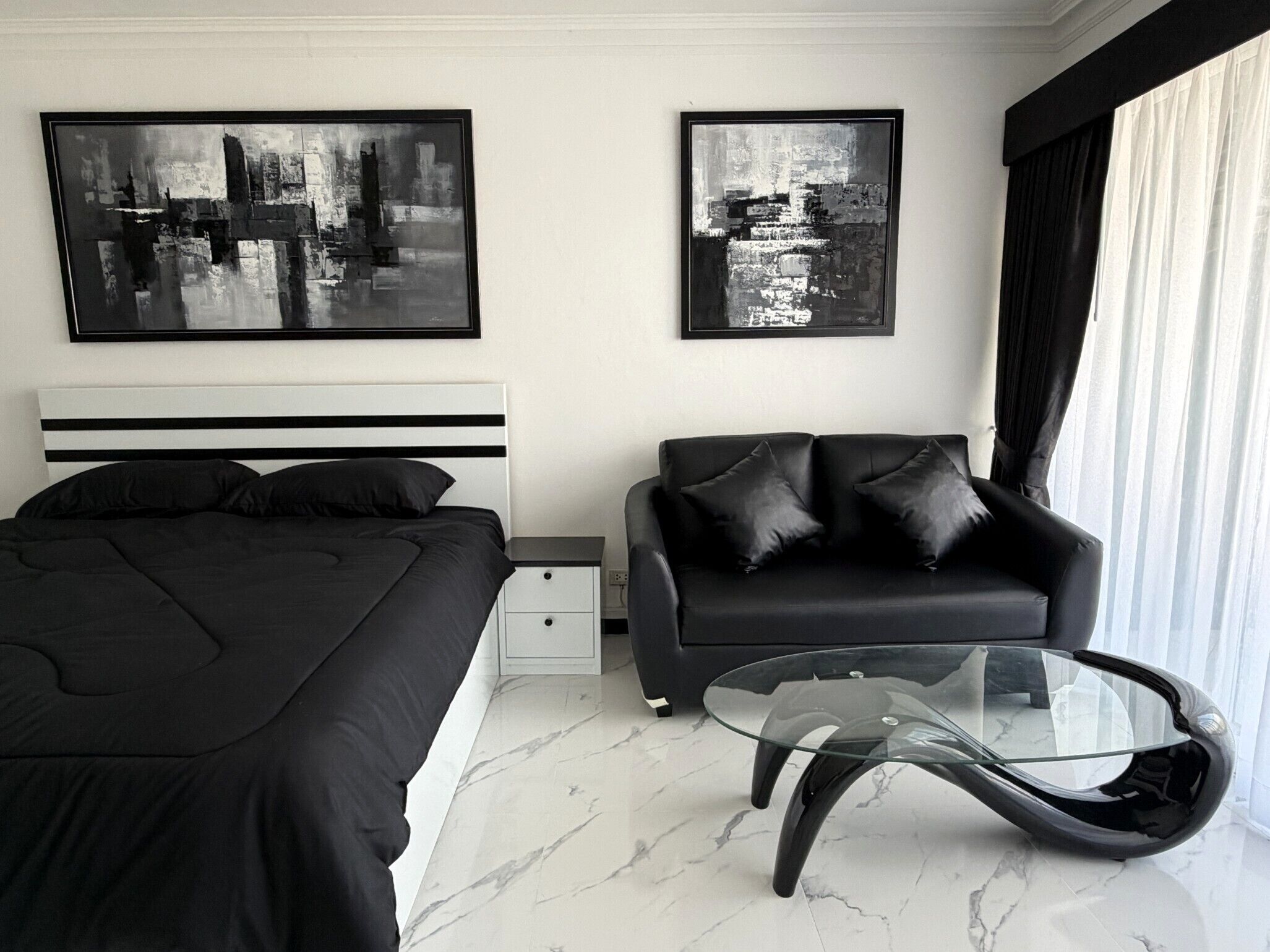 A modern bedroom with a bed, a sofa, a glass coffee table, and two abstract paintings on the wall.