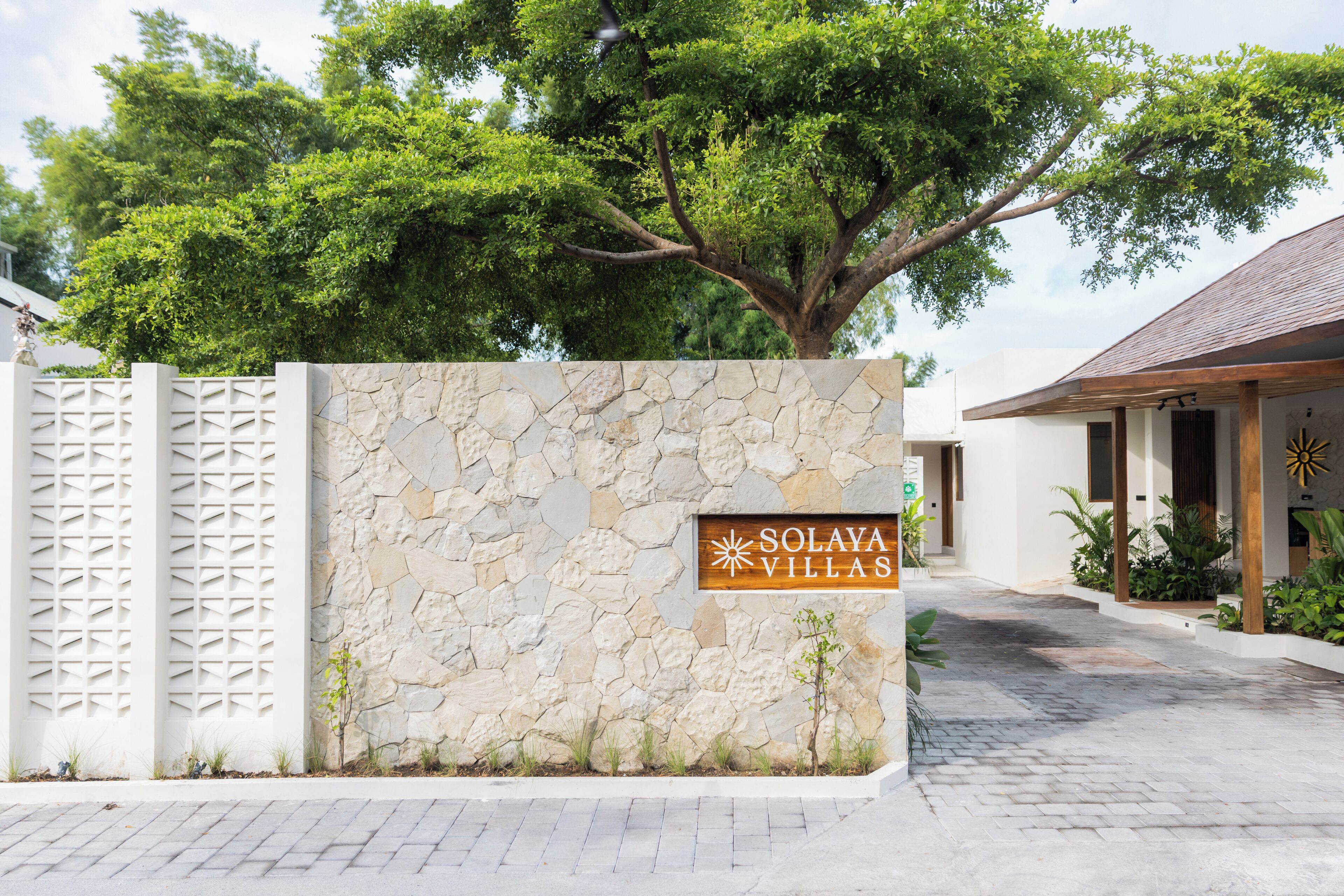 Un mur de pierre avec une enseigne indiquant « Solaya Villas », une allée pavée et un grand arbre.