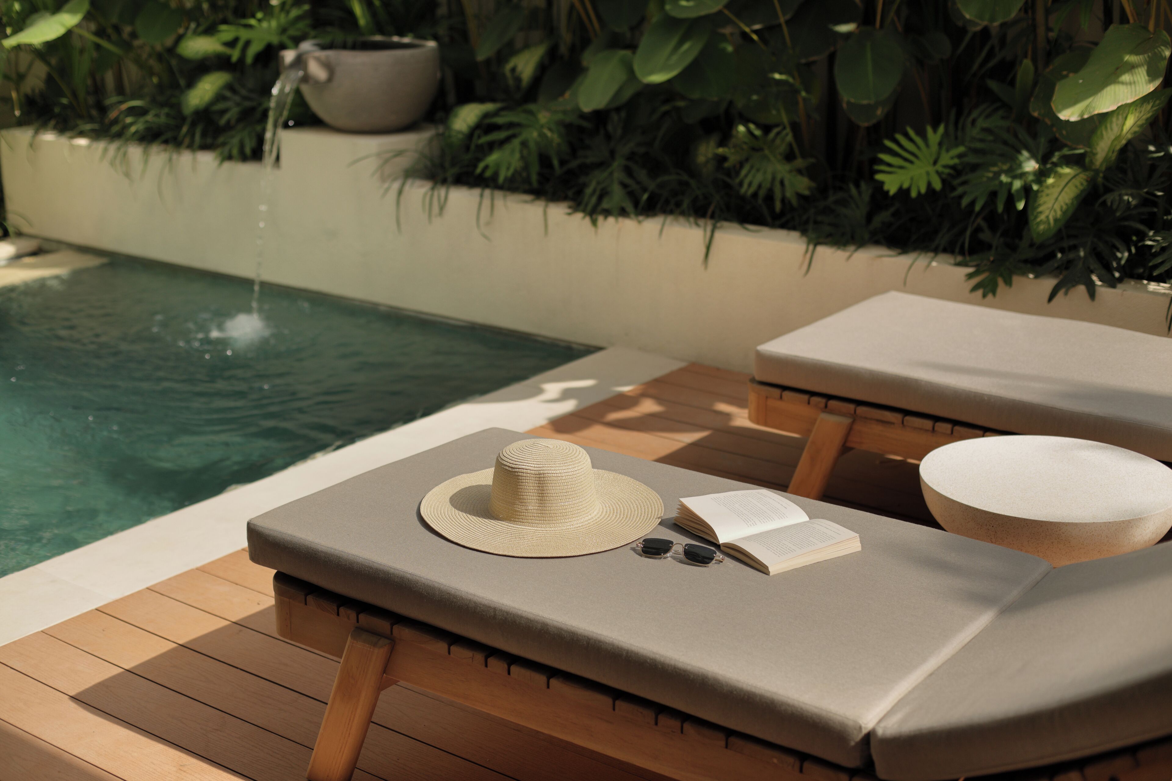 Un espace de détente au bord de la piscine, avec une terrasse en bois, un fauteuil, une petite table, un chapeau, des lunettes de soleil et un livre ouvert.