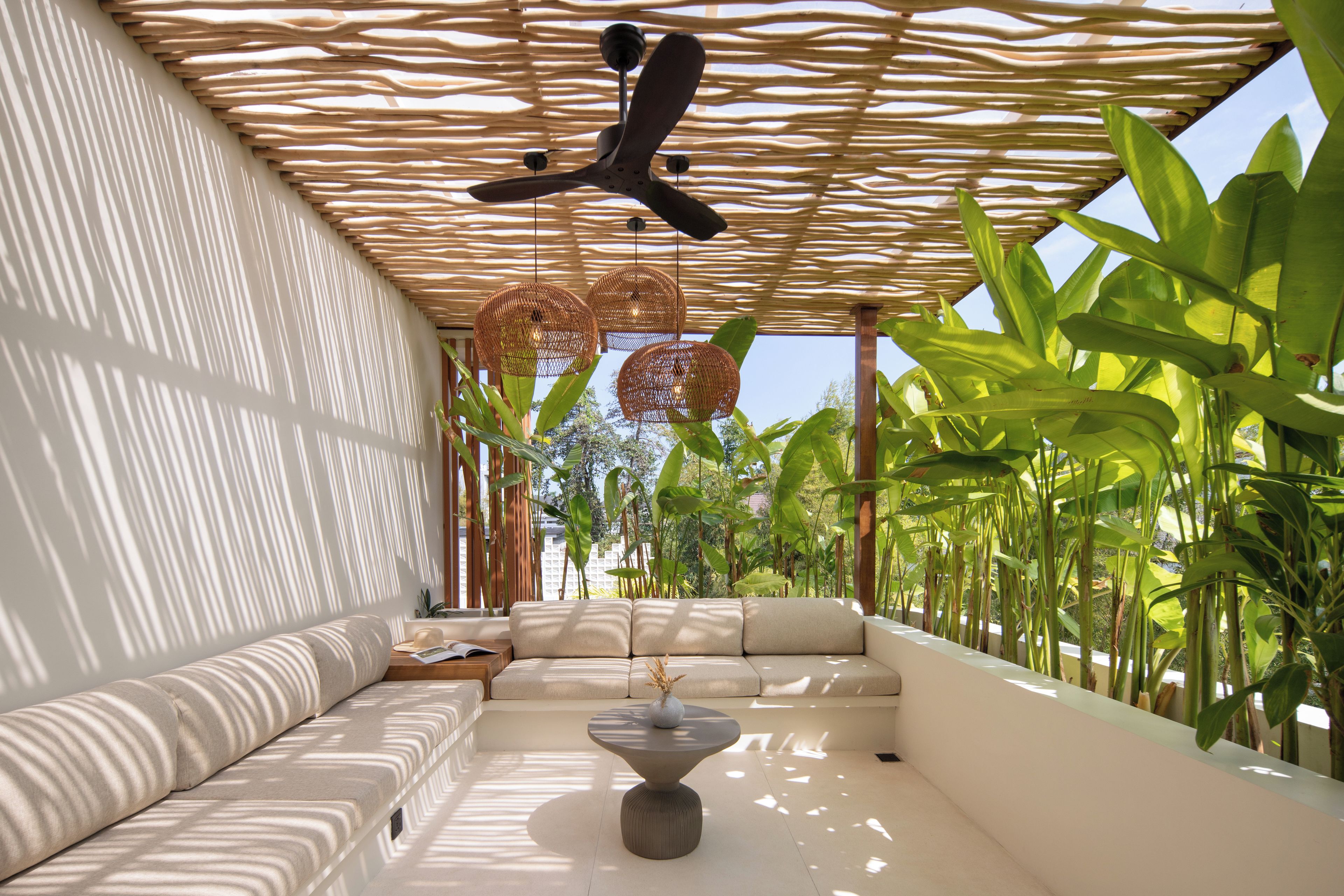 A modern outdoor seating area with a ceiling fan, woven light fixtures, and lush tropical plants.