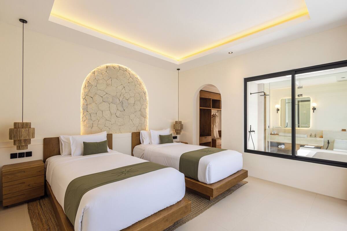 Une chambre d’hôtel moderne avec deux lits, un mur en pierre, un grand miroir et une table de chevet en bois.
