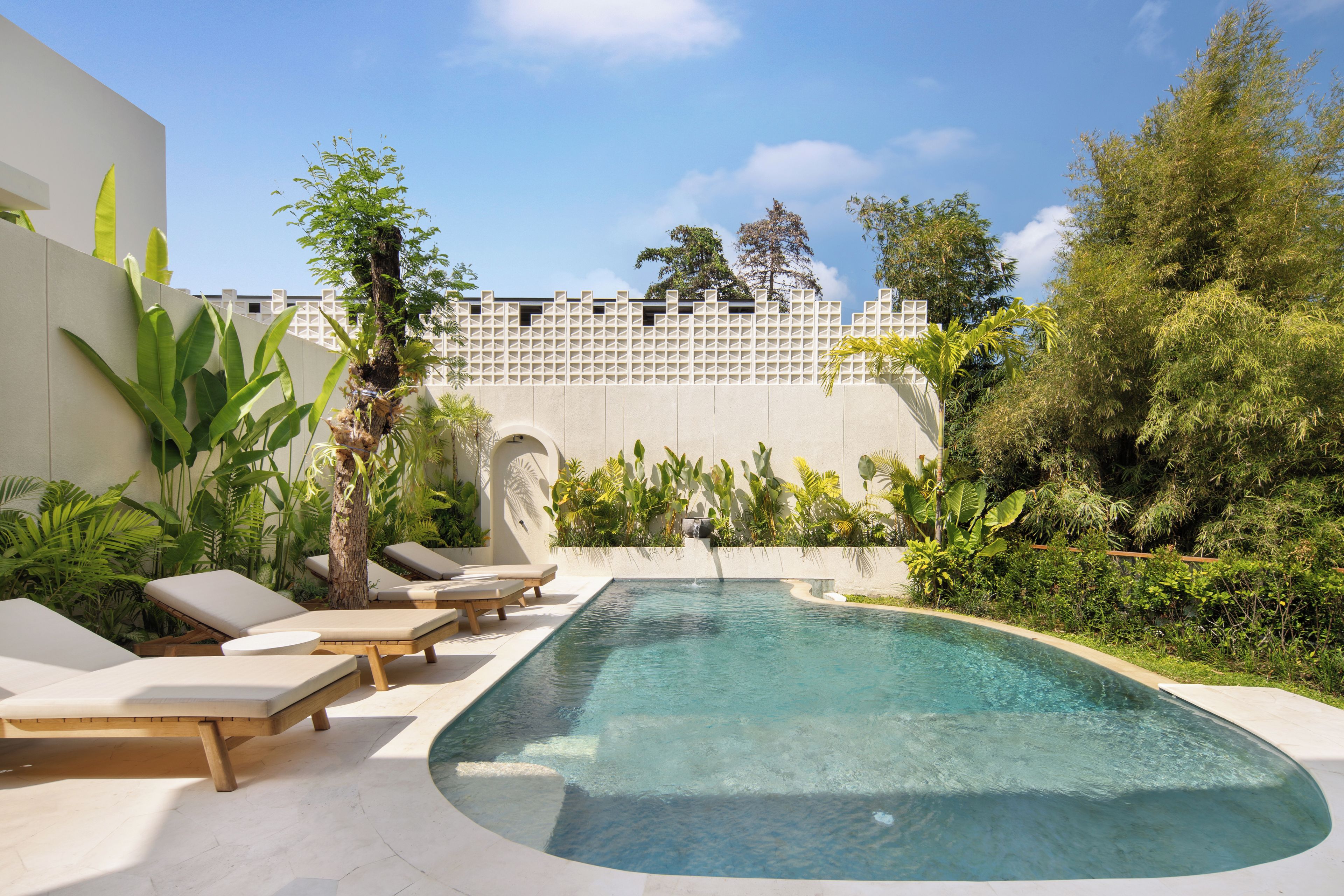 Una piscina con sillas de descanso, rodeada de plantas tropicales y una cerca blanca.