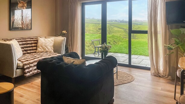 Un salón con sofá, un sillón negro, televisor y un amplio ventanal corredero que da a un balcón con vistas a un campo verde.