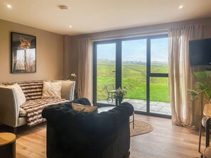 En stue med sofa, en sort lænestol, fjernsyn og en stor skydedør, der fører ud til en balkon med udsigt over et grønt område.
