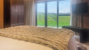 Un dormitorio con una cama grande, un televisor y vistas a un paisaje verde a través de un ventanal amplio.