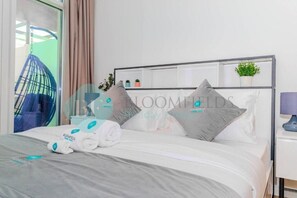 A neatly made bed with a grey bedspread and white pillows, a hanging chair, a small plant on a shelf, and a bedside table with a lamp. - Bloomfields Cozy 1BR In Oasis Masdar (Abu Dhabi)