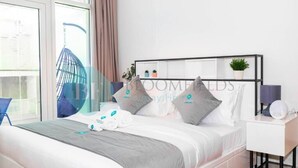 Una habitación de hotel moderna con una cama grande, una mesita de noche con lámpara, una silla y vistas de la ciudad a través de amplios ventanales.