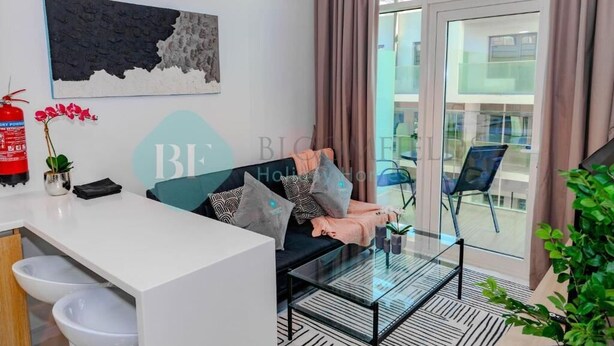 Una habitación de hotel moderna con un sofá, una mesa de centro de vidrio, un escritorio blanco y un ventanal con cortinas.