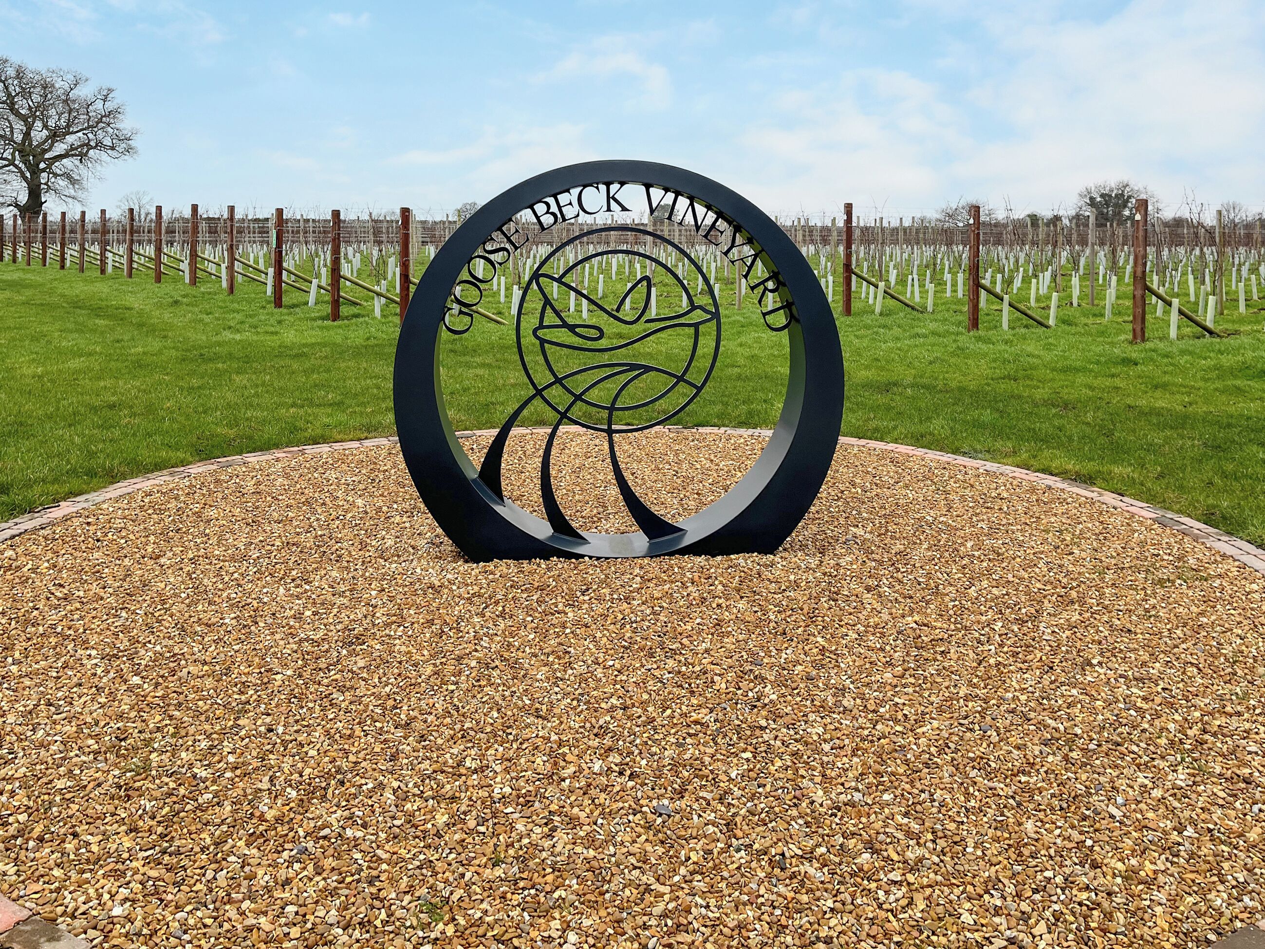 Un letrero metálico circular con el texto "CHOOSE BECK VINEYARD" rodeado por un camino de grava.