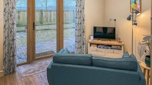 Ein Wohnzimmer mit Sofa, Fernseher und Bücherregal, großen Fenstern mit Vorhängen und Blick auf eine Holzterrasse.