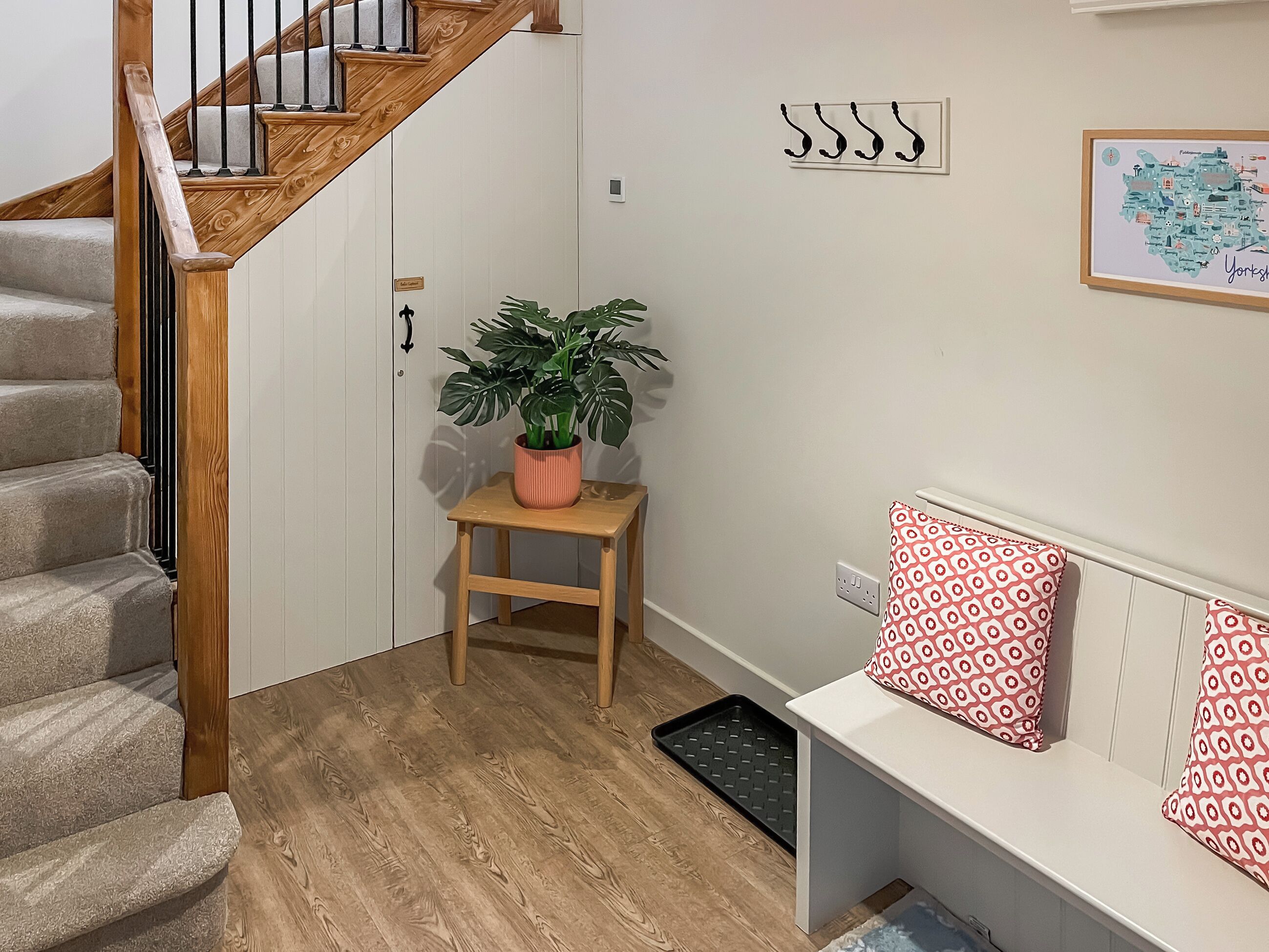 A staircase with wooden railings, a potted plant on a small stool, a white bench with patterned cushions, and a framed map on the wall.