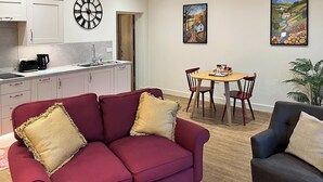 Ein modernes Wohnzimmer mit einem roten Sofa, einem dunklen Sessel und einem Holzes Esstisch mit vier Stühlen. Es gibt eine Küchenzeile mit weißen Schränken und einer Uhr an der Wand.