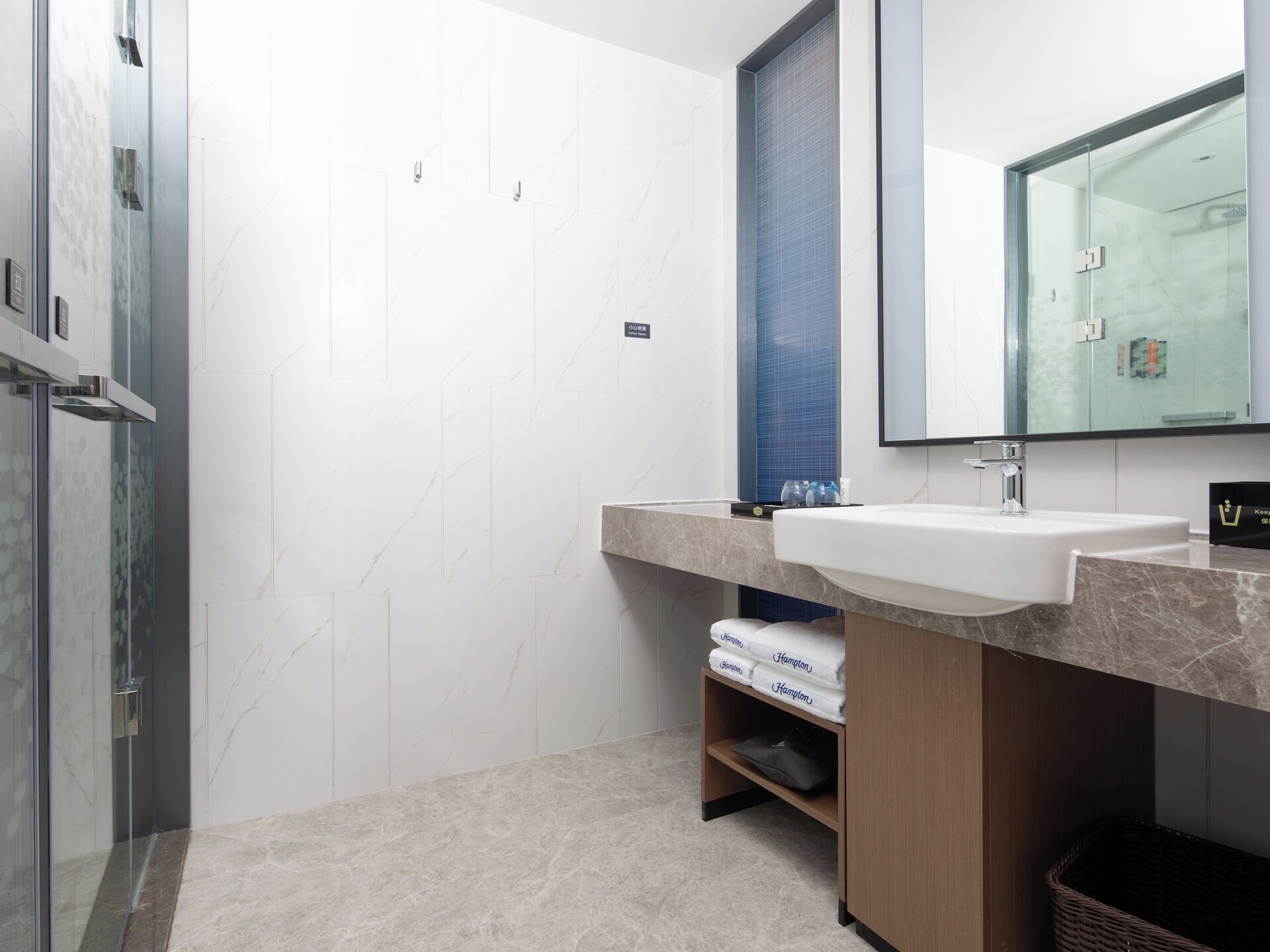A modern bathroom with a glass shower enclosure, a white sink, and a mirror above it. There are towels and a basket on the counter.