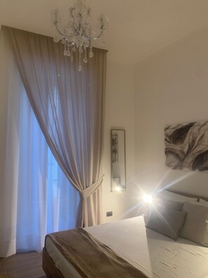 A bedroom with a bed, a chandelier, a large window with curtains, a painting on the wall, and a mirror. - AR.b&b in the heart of Naples (Napoli)