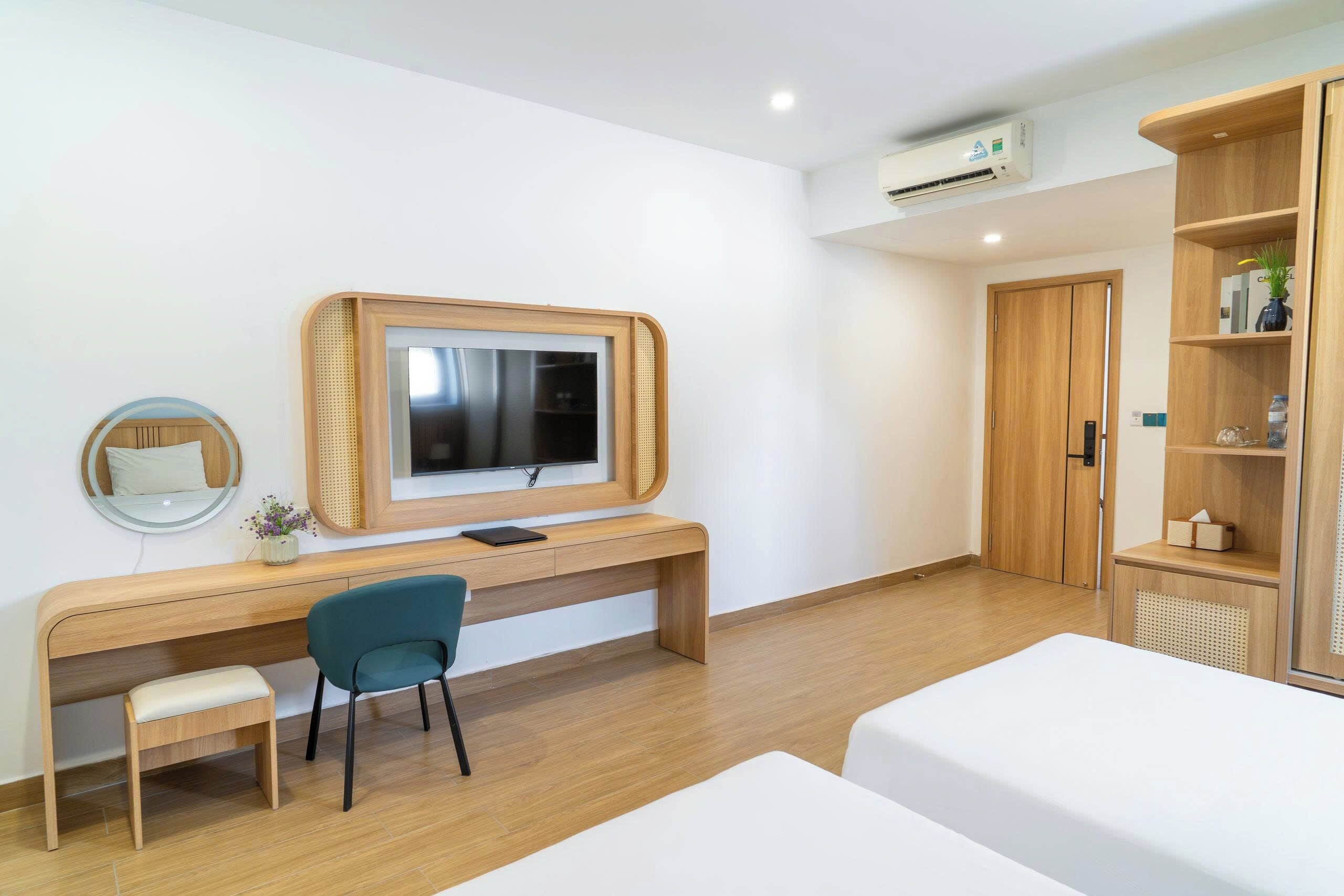 A modern hotel room with a wooden desk, a television, a small chair, a bed, and a wardrobe.