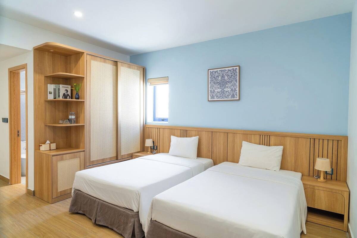 A hotel room with two beds, wooden headboards, a large window with blinds, a wall-mounted picture, and a wooden wardrobe with shelves.