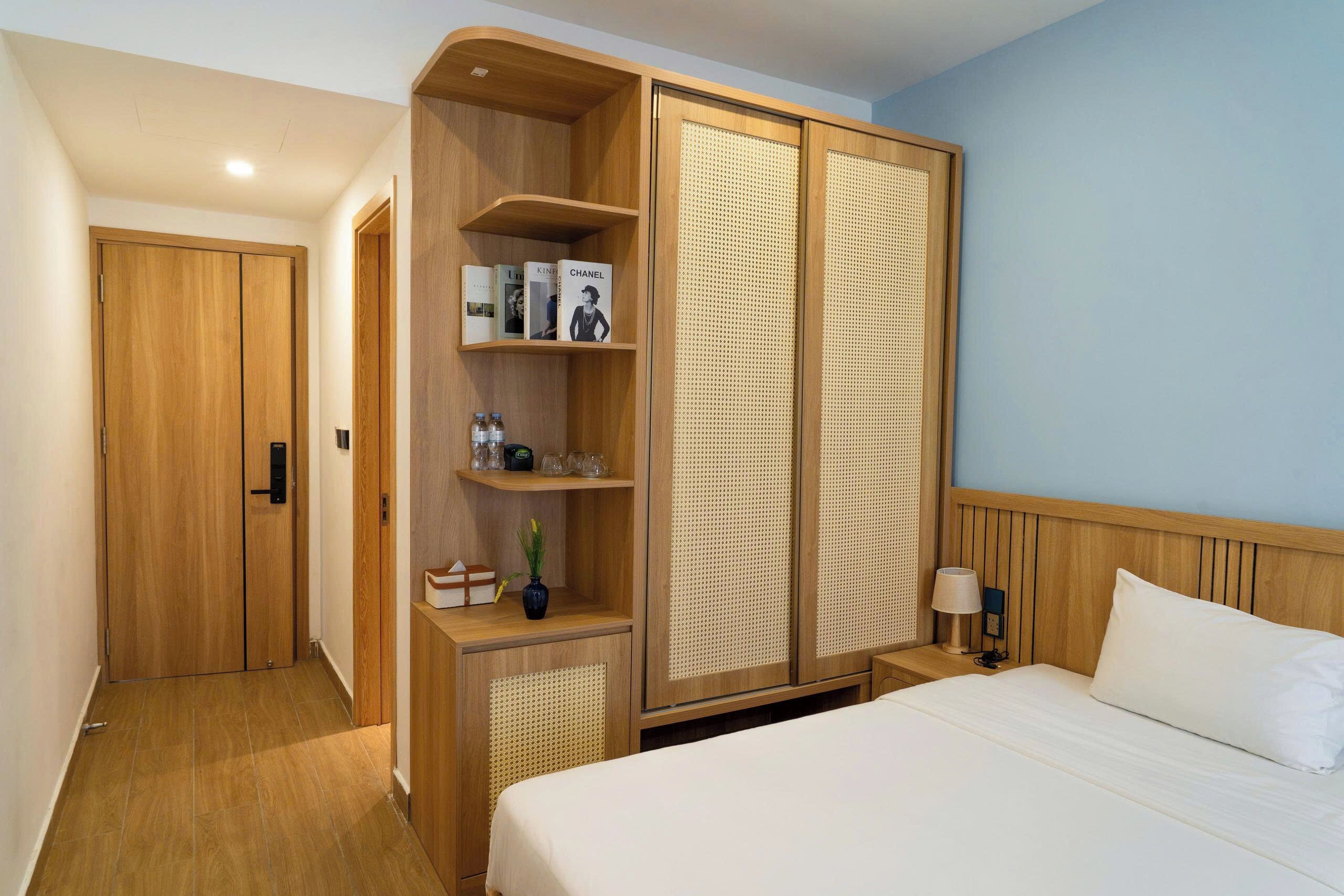 A hotel room with a wooden wardrobe, a bed with white linens, a nightstand with a lamp, and a small shelf with books and a vase.