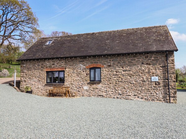 A stone building with a gravel driveway, two windows, and a small bench outside. - The Granary (Welshpool)