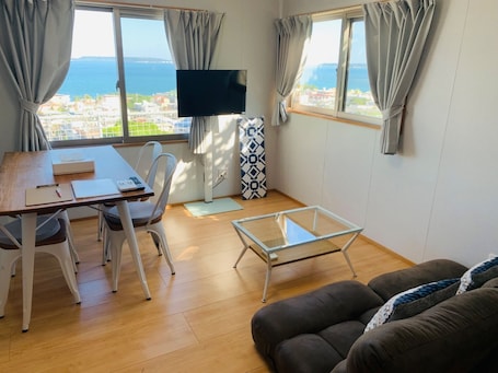 A modern living room with a wooden dining table, white chairs, a glass coffee table, and a brown sofa. There is a flat-screen TV mounted on the wall, and large windows with curtains offering a view of the sea and buildings.