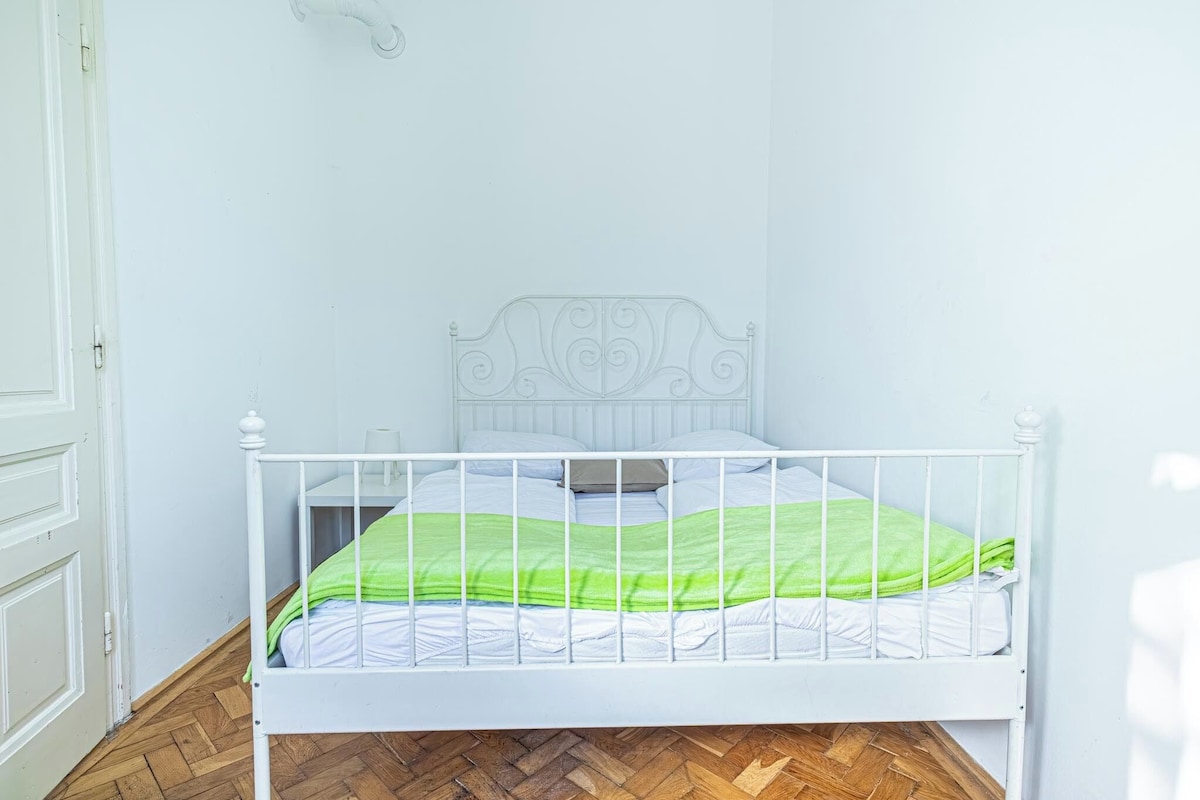 Ein weißes Metallbett mit grüner Decke in einem Zimmer mit weißen Wänden und Holzboden.