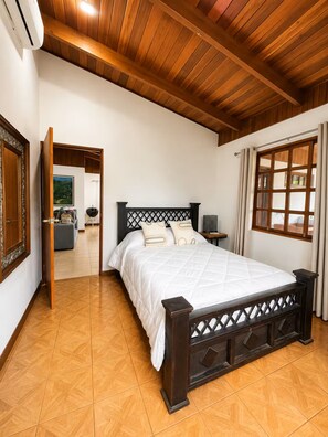 Un dormitorio con techo de madera, una cama con cabecera, una ventana con cortinas y una puerta que da a otra habitación.