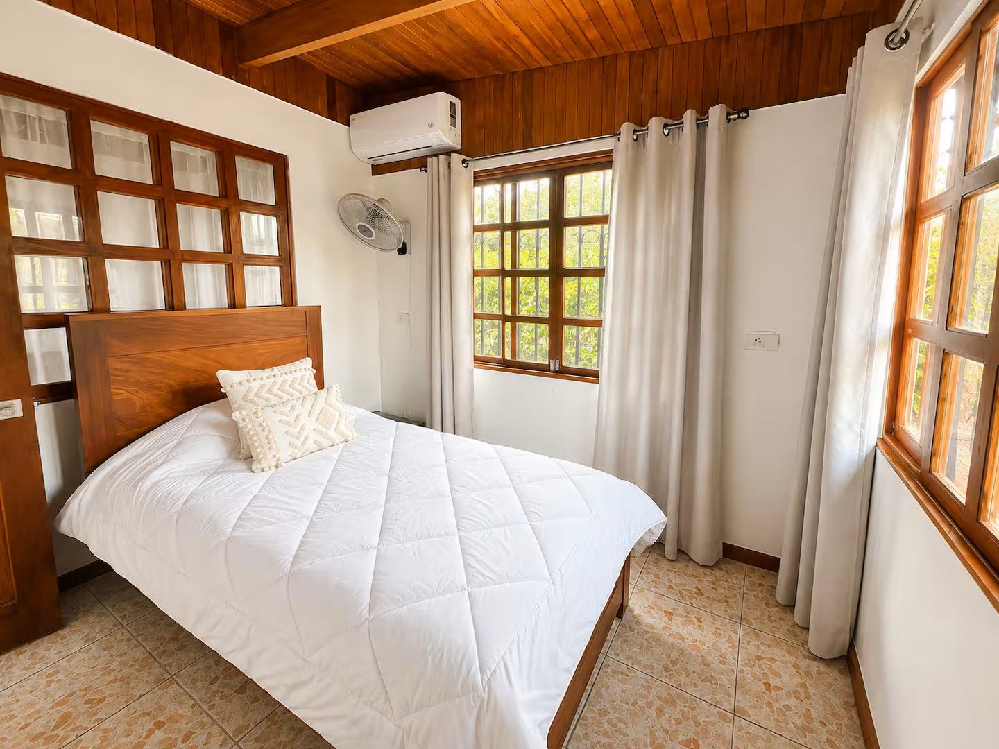 A bedroom with a wooden headboard, a bed with white bedding, a window with curtains, and an air conditioning unit.