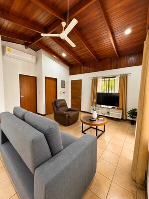 A spacious living room with a ceiling fan, a grey sofa, a leather chair, a coffee table, and a television. - Charming 3-bedroom House Rental in Paquera, Costa Rica (Paquera)