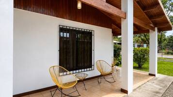A covered outdoor seating area with two chairs and a window with metal bars.