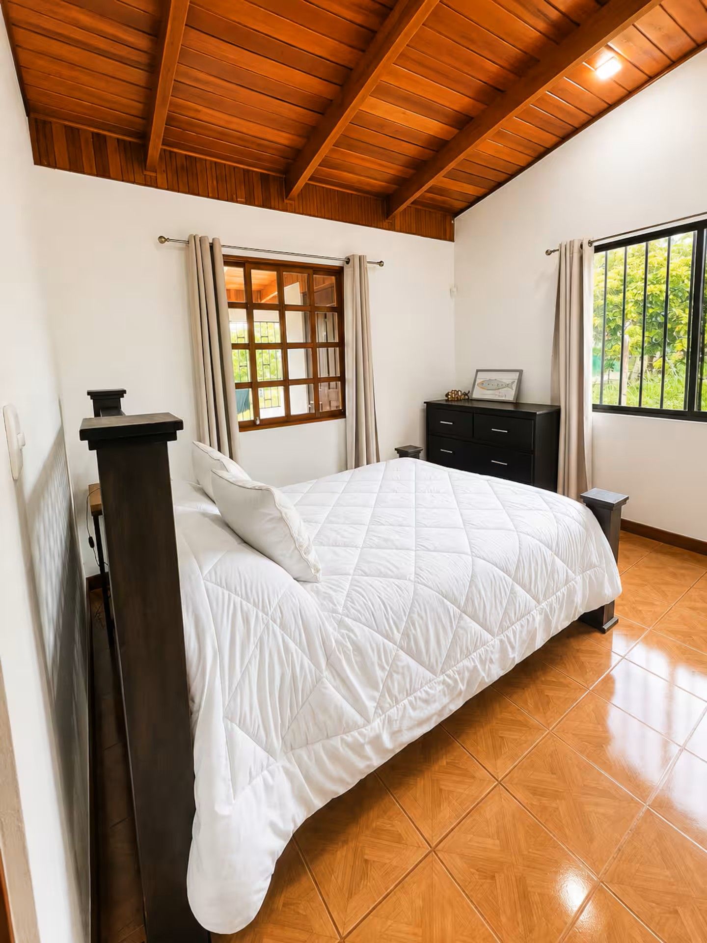 A bedroom with a wooden ceiling, a bed with a white quilt, a dresser, and a window with curtains.