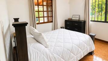 A bedroom with a wooden ceiling, a bed with a white quilt, a dresser, and a window with curtains.
