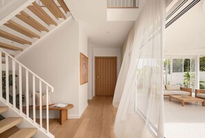 Eine Treppe mit Holzbeschlägen und weißen Geländern, eine Holztür sowie ein Blick auf einen Außenbereich mit Sitzgelegenheiten durch transparente Vorhänge.