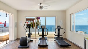 Ein Fitnessraum mit Laufbändern, einem Hometrainer und einem Rudergerät, mit Blick auf den Ozean durch große Fenster.