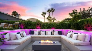 Zona relax esterna moderna con un elemento decorativo a fuoco, circondata da vegetazione rigogliosa e palme, sotto un cielo crepuscolare.