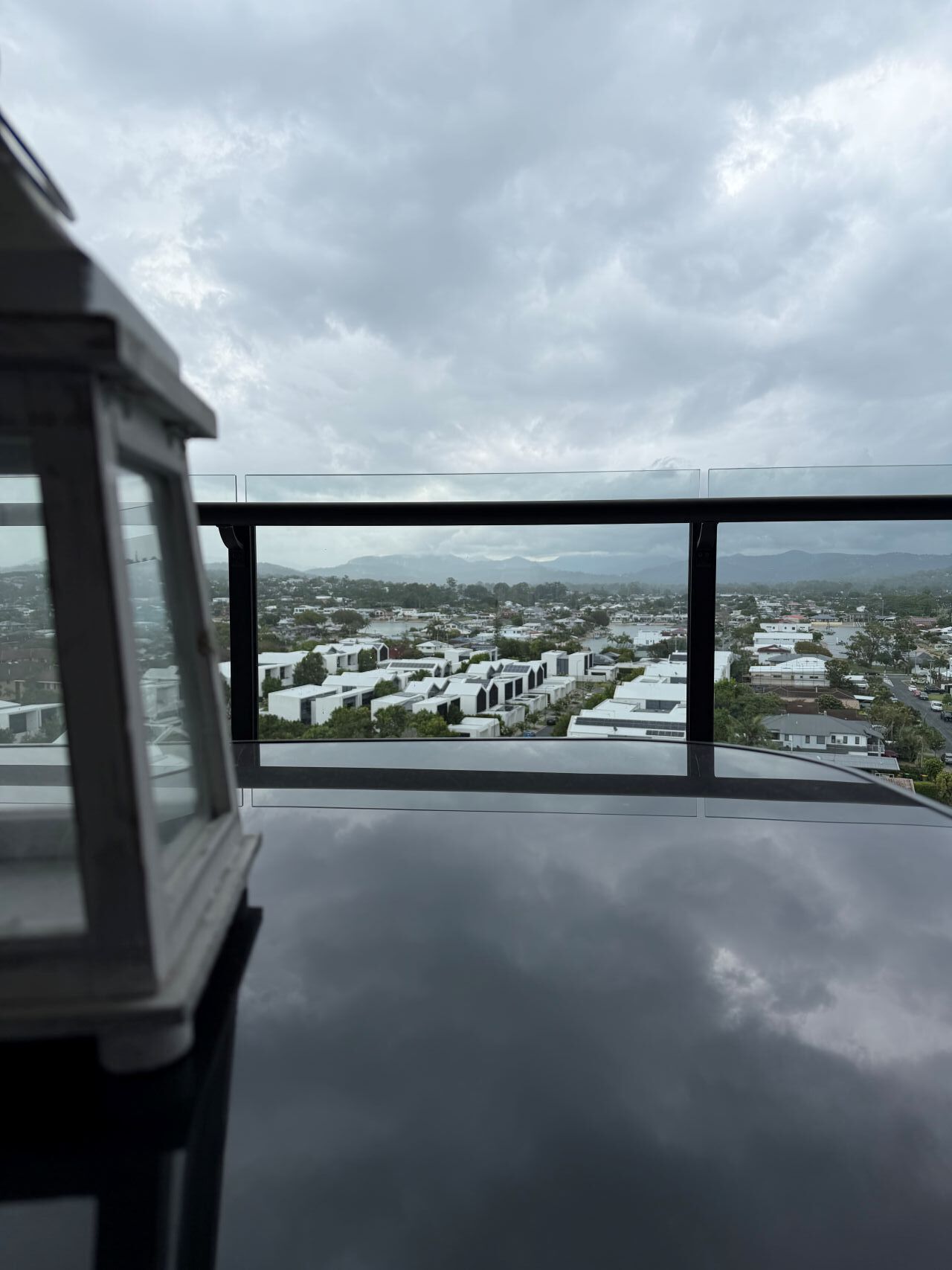 A view from a high-rise building with a glass railing, overlooking a cityscape with modern buildings and a cloudy sky.