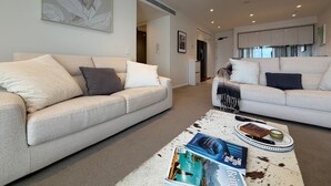 Een moderne woonkamer met twee beige banken, een salontafel met tijdschriften, en een keuken op de achtergrond.