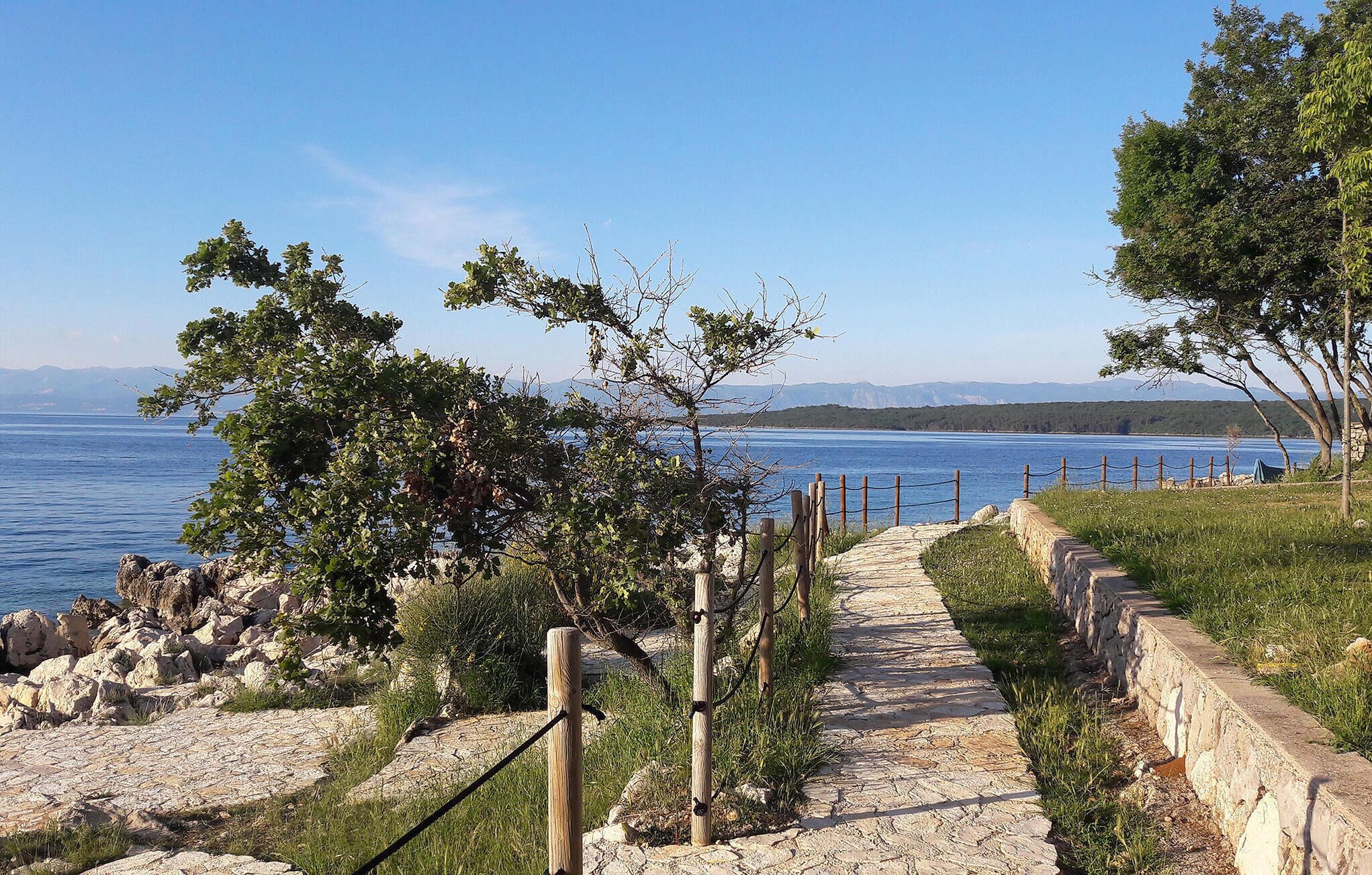 Un sentier de pierres menant à un plan d’eau, bordé d’arbres et d’un mur de pierres.