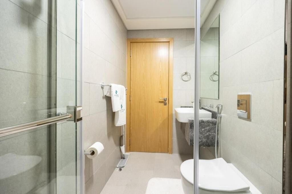 A narrow bathroom with a glass shower enclosure, a wooden door, a white sink, and a toilet.