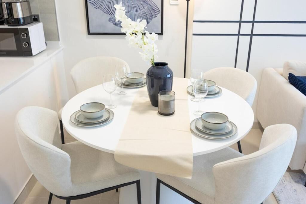 Uma mesa de jantar redonda com cadeiras brancas, um vaso com flores brancas e uma cafeteira à esquerda.