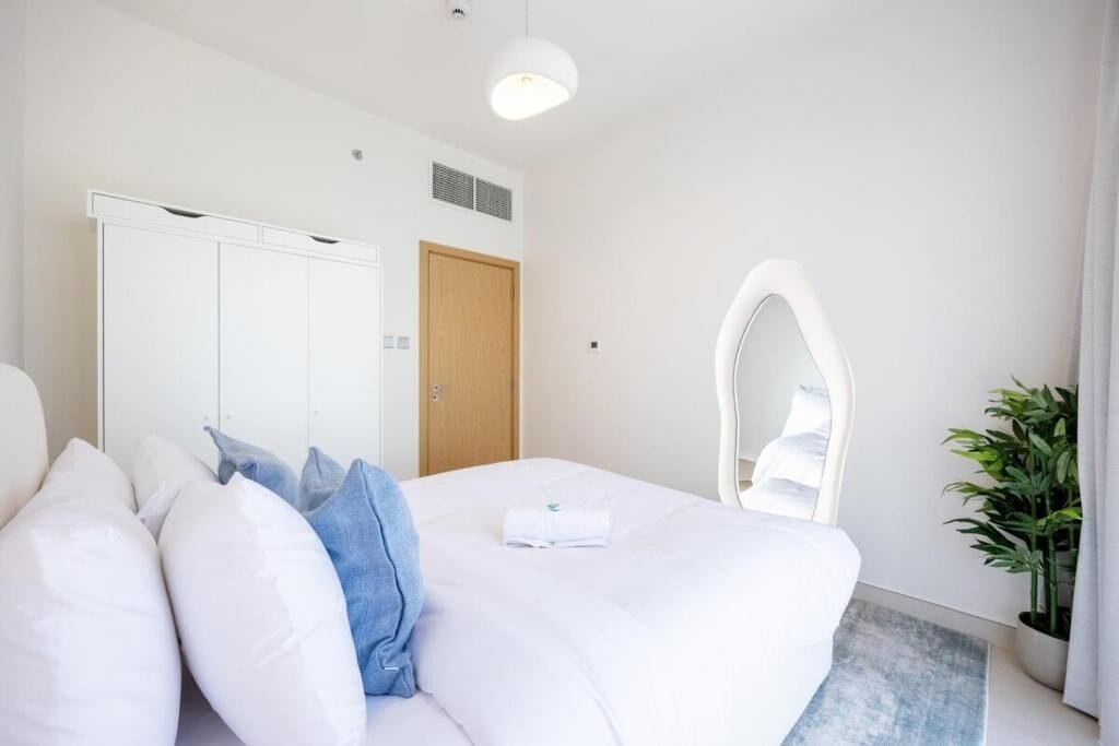 Une chambre d’hôtel moderne avec un grand lit, du linge de lit blanc, une porte en bois, une armoire blanche, un miroir et une plante en pot.